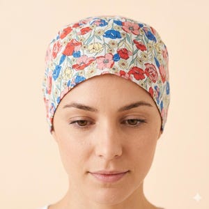 May include: A scrub cap featuring a floral pattern with red, blue, and yellow flowers on a white background. The cap has a rounded crown and a fitted design that covers the top of the head, showcasing a vibrant botanical print with green stems.