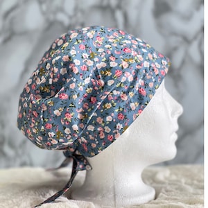 May include: A floral patterned scrub cap in shades of blue, pink, and green. The cap is tied at the back with matching fabric ties. The cap is displayed on a white mannequin head.