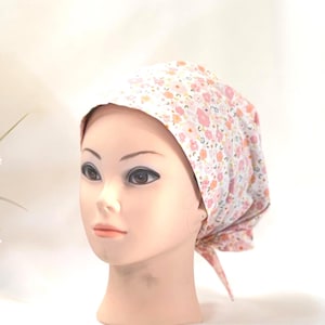 May include: A headscarf with a floral pattern in pink, peach, and white. The fabric is adorned with small flowers and leaves. The headscarf is tied at the back.