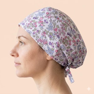 May include: A scrub cap featuring a light lavender background with a dense floral pattern in shades of pink, purple, blue, and green. The cap has a rounded crown and includes two fabric ties at the back to secure the fit around the head.