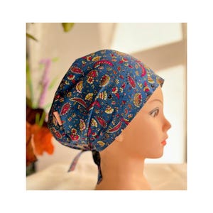 Op de afbeelding: Een blauwe chirurgische scrubcap met een kleurrijk bloemenpatroon. De cap heeft een strik aan de achterkant.