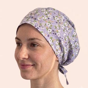 May include: A lavender scrub cap featuring a repeating floral pattern of white blossoms with yellow centres and small dark purple flowers. The fabric head covering has a rounded crown and ties at the back to secure the fit around the head.