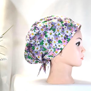 Può includere: Un cappello chirurgico a fantasia floreale con stampa a margherita bianca, gialla, viola e rosa su sfondo lilla chiaro. Il cappello è legato sul retro ed è esposto su una testa di manichino.