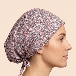 May include: A scrub cap featuring a repeating floral pattern with small white flowers, dark blue centres, and green leaves on a dusty pink background. The cap has a rounded crown and a tie closure at the nape of the neck to secure the fit.