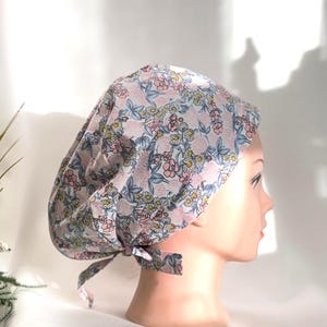 Puede incluir: Un gorro quirúrgico con estampado floral, con un fondo rosa claro y un patrón repetido de flores rosas y amarillas y hojas azules. El gorro se ata en la parte posterior con una corbata de tela a juego.