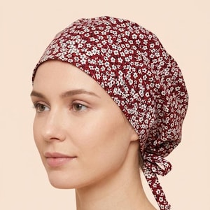 May include: A maroon scrub cap featuring a dense pattern of small white flowers with yellow centers. The head covering has a rounded crown and includes two fabric ties at the back that are knotted to secure the fit around the head.