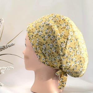 Peut inclure: Un couvre-chef à motifs floraux jaunes. Le tissu présente un motif répété de petites fleurs blanches avec des accents verts et marron. Le couvre-chef est noué à la nuque.