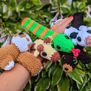 May include: A selection of handmade crochet toys, including a brown bear, a giraffe, a snake, a cow, and a dachshund. The toys are in various colours, such as brown, green, white, and black. The snake is striped with green and orange.