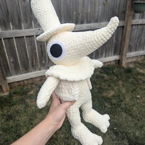 Op de afbeelding: Een crèmekleurige gehaakte knuffel met een hoge hoed, een lange puntige neus en een groot rond zwart oog. Het figuur heeft een geplooide kraag, een lichaam van getextureerd garen en voeten met drie tenen, gemaakt van zacht, dik garen.