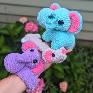 May include: Three crocheted elephant toys in the image. One is light blue with pink ears, another is purple with pink ears, and the third is gray with pink ears. The elephants have large black eyes and are handmade.