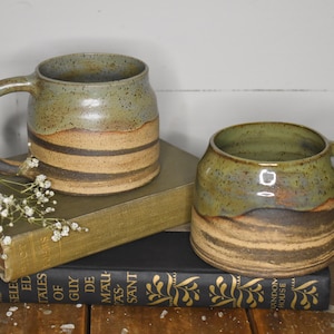 May include: Two handmade ceramic mugs with a green glaze and brown stripes. The mugs are sitting on top of a stack of books.