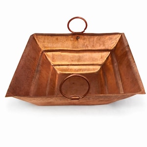 May include: A square copper tray with a hammered texture and a metallic finish. The tray features a stepped, tiered interior design and two circular copper handles attached to opposite sides of the rim, providing a functional grip for the vessel.