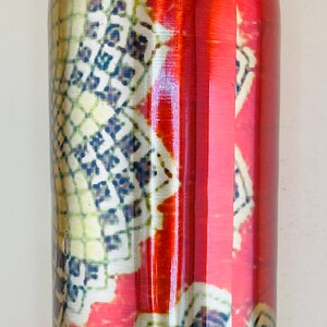 May include: A tall, cylindrical water bottle with a decorative pattern. The bottle has a red background with a repeating pattern of white, yellow, and blue geometric shapes. The lid is brown and gold.