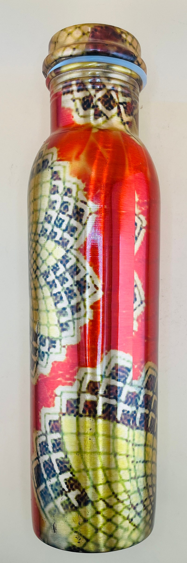 May include: A tall, cylindrical water bottle with a decorative pattern. The bottle has a red background with a repeating pattern of white, yellow, and blue geometric shapes. The lid is brown and gold.