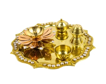 May include: A golden-colored puja thali set with a decorative edge, featuring a central lotus-shaped lamp, small bowls, and a covered container. The thali is adorned with clear, sparkling stones around the edge.