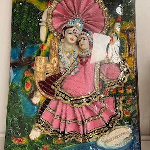May include: A 3D wall art piece depicting a Hindu couple, Radha and Krishna, in a colorful, ornate setting. The couple is dressed in pink and gold, with intricate details and embellishments. The artwork is framed and features a green background with a blue river and a white swan.