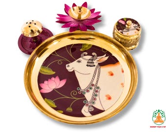 Può includere: Un set puja decorativo con un piatto bordato d'oro con un'illustrazione di mucca, un piccolo contenitore dorato, un bruciatore d'incenso dorato e una lampada a olio a forma di loto rosa. Il set è progettato per cerimonie religiose.