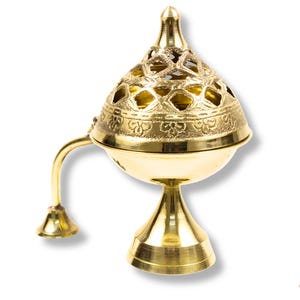 May include: A gold-coloured incense burner with a decorative, pierced lid and a curved handle. The burner has a round base and a conical top, with intricate patterns etched into the metal. The overall design is ornate and traditional.