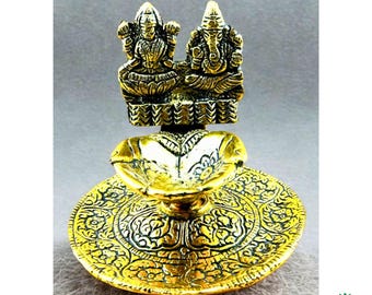 May include: A gold-colored decorative oil lamp featuring detailed carvings. The lamp has a round base with an intricate floral design, a central bowl, and a top section with two figures. The figures are seated and appear to be religious deities.