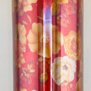 May include: A tall, cylindrical water bottle with a floral design. The bottle has a red background with gold and cream-colored flowers. The lid is a light blue color.