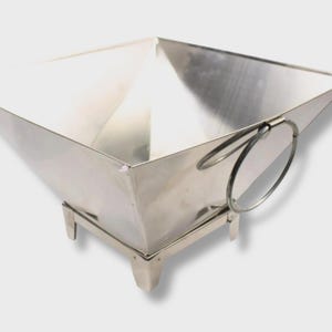 May include: A stainless steel square container with a flared design and small feet. The container has a shiny, reflective surface and two circular handles on one side. The container is designed to hold items.