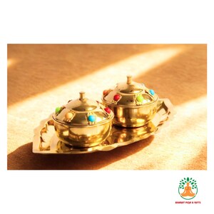 May include: Two gold-colored, lidded bowls with colorful gemstone accents, resting on a matching tray. The bowls have a rounded shape and small handles. The tray has a decorative, scalloped edge. The items are displayed on a light brown surface.