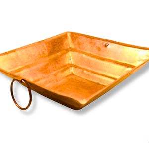 May include: A square, copper-colored metal tray with a rounded handle. The tray has a shallow, open design with slightly raised edges. The copper finish gives the tray a warm, inviting appearance. The tray is likely used for decorative or serving purposes.