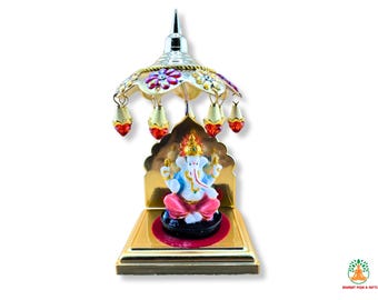 May include: An ornate Ganesh idol, with a white and blue figure seated on a black base, within a gold-coloured frame. The idol is topped with a silver canopy with floral and hanging accents. The base is gold, and the design is detailed.