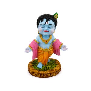 Könnte beinhalten: Eine Babyfigur mit hellblauer Haut, schwarzem Haar und einer Pfauenfeder als Accessoire. Die Figur trägt eine goldene Halskette, goldene Armreifen, einen gelben Dhoti und einen rosa Schal. Sie steht auf einem strukturierten, runden Sockel.