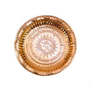May include: A round, gold-colored metal plate with embossed details. The center features the Om symbol and radiating lines, surrounded by text. The plate has a textured rim and is set against a white background.
