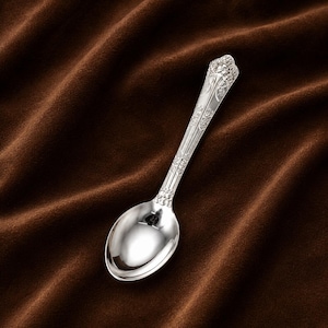 May include: A silver-toned metal spoon featuring an ornate, embossed floral pattern along the handle. The handle tapers toward the top, which is rounded and decorated with intricate relief details, while the bowl of the spoon is smooth and reflective.
