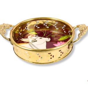 May include: A decorative gold-colored bowl with ornate handles. The bowl's interior features a painted design of a white cow with a floral and leaf pattern. The bowl has a maroon background and is likely used for decorative purposes.