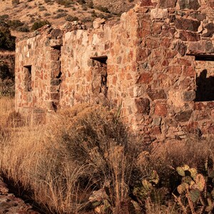 May include: A weathered stone building ruin with exposed walls and empty window frames, constructed from irregular brown and tan rocks. The structure is surrounded by dry golden grasses and prickly pear cacti in a desert landscape under sunlight.