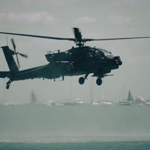 Könnte beinhalten: Ein dunkel gefärbter militärischer Kampfhubschrauber im Flug, ausgestattet mit einem markanten Hauptrotorsystem, einem Heckrotor und einer Cockpithaube. Das Fluggerät befindet sich über einem Gewässer, auf dem mehrere kleine Boote zu sehen sind.