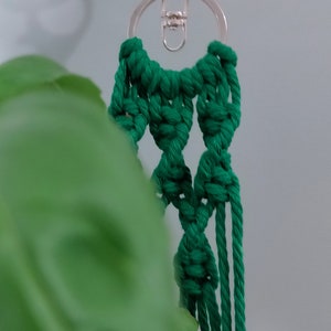 May include: A green macrame keychain with a silver key ring and a silver clasp. The keychain is hanging from a green leaf.