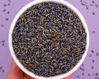 May include: A white ceramic bowl filled with dried lavender buds. The small, elongated flower buds are a mix of deep purple, blue, and muted green tones, creating a dense, textured surface within the circular container.