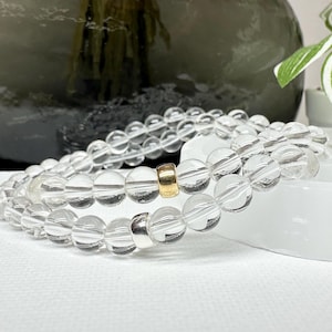 Könnte beinhalten: Zwei Armbänder aus klaren Quarz-Kristallperlen. Jedes Armband hat runde, transparente Perlen, die zusammen aufgefädelt sind. Ein Armband hat eine goldfarbene Abstandshülse, das andere eine silberfarbene. Die Armbänder werden auf einer weißen Oberfläche präsentiert.