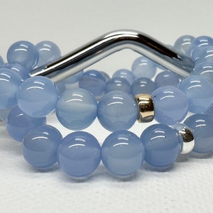May include: Two light blue beaded bracelets. Each bracelet is made of large, round, polished beads. One bracelet has a gold-coloured spacer bead, and the other has a silver-coloured spacer bead. The bracelets are displayed on a white surface.