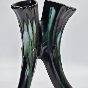 May include: A black ceramic vase with a unique, organic shape. The vase features two tall, fluted openings connected by a curved base. Green and blue glaze accents run down the sides, creating a striking contrast against the black.