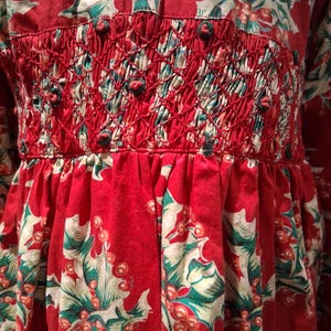 May include: A red dress with a festive holly print featuring green leaves and red berries. The bodice has a smocked design with red stitching and small, decorative knots. The skirt is gathered, creating a flowing silhouette.