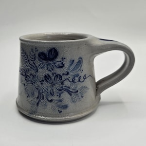 May include: A gray ceramic mug with a large handle. The mug is decorated with a blue floral pattern. The flowers and leaves are hand-painted. The mug has a slightly textured surface.