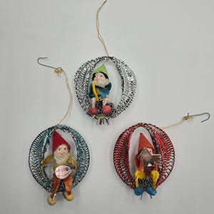 May include: Three vintage Christmas ornaments featuring gnome figures. Each gnome is inside a wireframe sphere. One gnome wears a red hat and plays a mandolin. Another wears a green hat and holds a horn. The third gnome wears a red hat and reads a book.