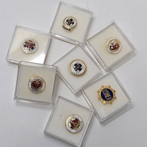 May include: Seven small, clear plastic boxes each containing a different pin with a white background and gold trim. The pins feature various medical symbols and text, including "Respiratory Therapy", "Nursing Assistant", "Usher", "Medical", "Volunteer", and "First Aid".