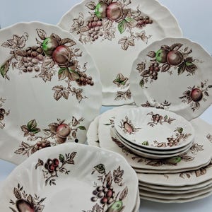 May include: A collection of vintage white ceramic dishware with a fruit and grape design. The set includes plates, bowls, and saucers. The fruit is depicted in shades of green, red, and brown, with green leaves and brown vines.