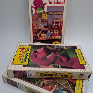 May include: Three vintage Barney & Friends VHS tapes. The top tape is titled "Barney Goes To School" with a picture of Barney. The other two tapes are titled "Barney & Friends" with images of Barney and children. The tapes are in their original packaging.