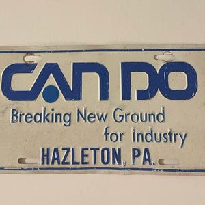 Puede incluir: Placa de matrícula rectangular con las palabras "CAN DO" en azul, encima del texto "Breaking New Ground for Industry" y "HAZLETON, PA." La placa tiene un fondo blanco y un borde azul.