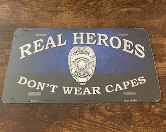 May include: A blue and black metal sign with the text "REAL HEROES DON'T WEAR CAPES" and a silver police badge with the word "OFFICER" and "POLICE" on it. The sign is made in the USA.