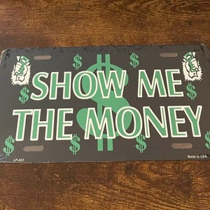 以下が含まれることがあります： 黒い金属製のナンバープレートに、緑色の文字で「SHOW ME THE MONEY」と書かれています。プレートは、ドル記号と、ドル札を持った手のスタイリッシュな画像で飾られています。