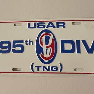 Może przedstawiać: Biała tablica rejestracyjna z czerwoną obwódką i napisem "USAR", "95th DIV" i "(TNG)" w kolorze niebieskim. Centralny emblemat przedstawia niebieski owal z czerwono-białym wzorem. Tablica ma cztery otwory montażowe.