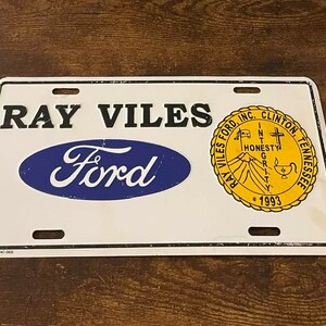 May include: White metal license plate with blue and black text reading "RAY VILES Ford" and a yellow circular seal with the text "RAY VILES FORD, INC. CLINTON, TENNESSEE" and "HONESTY INTEGRITY GRIT" around a mountain range with a cross and a candle.